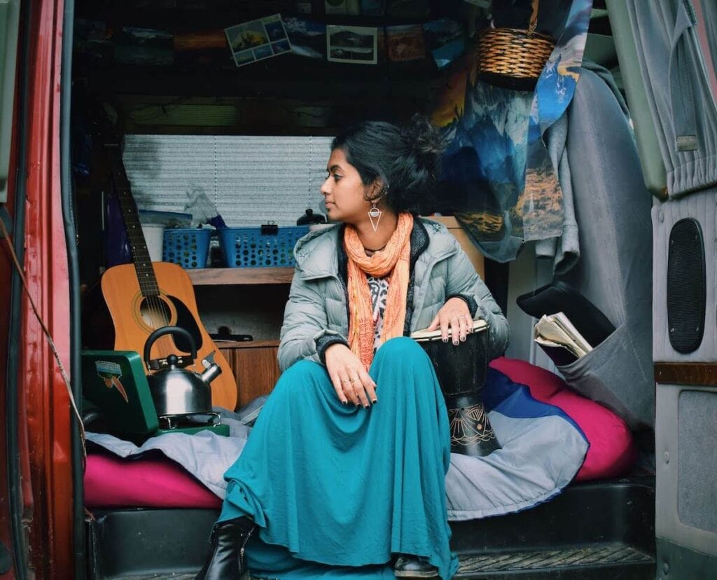 A woman sits in the doorway of her campervan as she looks away and has her hand rested on a small hand drum