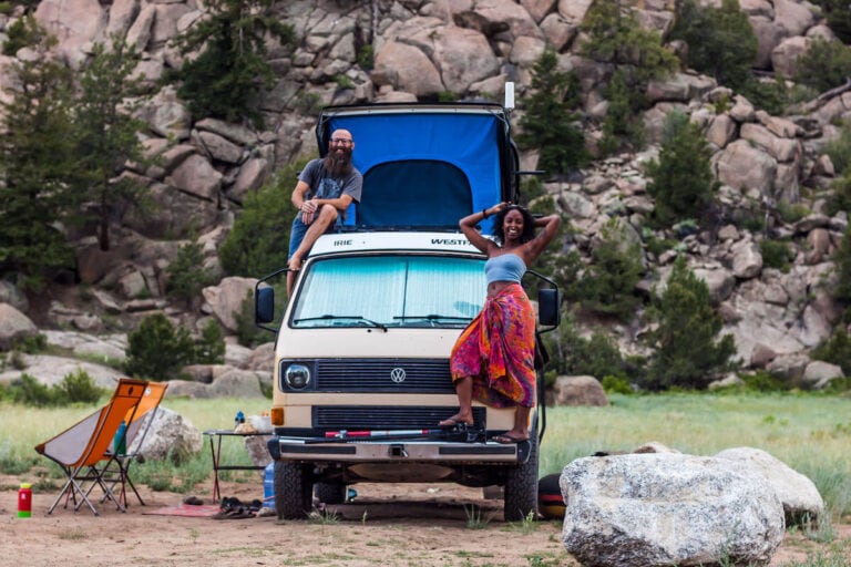 a man and woman standing on their yellow VW bus with a pop top