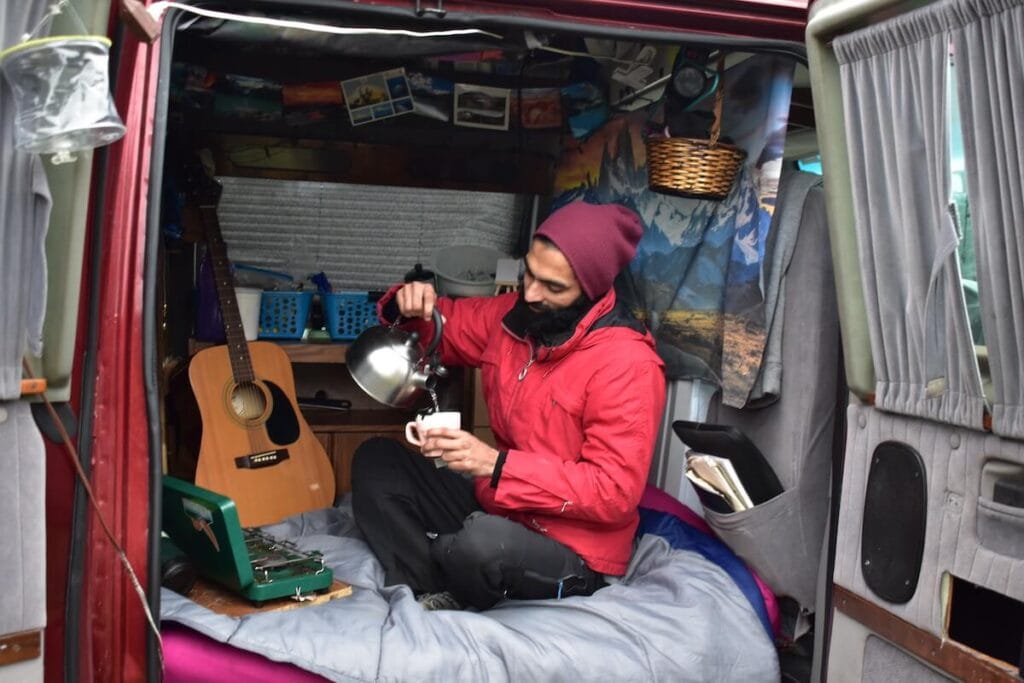 A man wearing a red jacket and beanie pouring water from a kettle into a mug sitting on a bed with the doors of his van open
