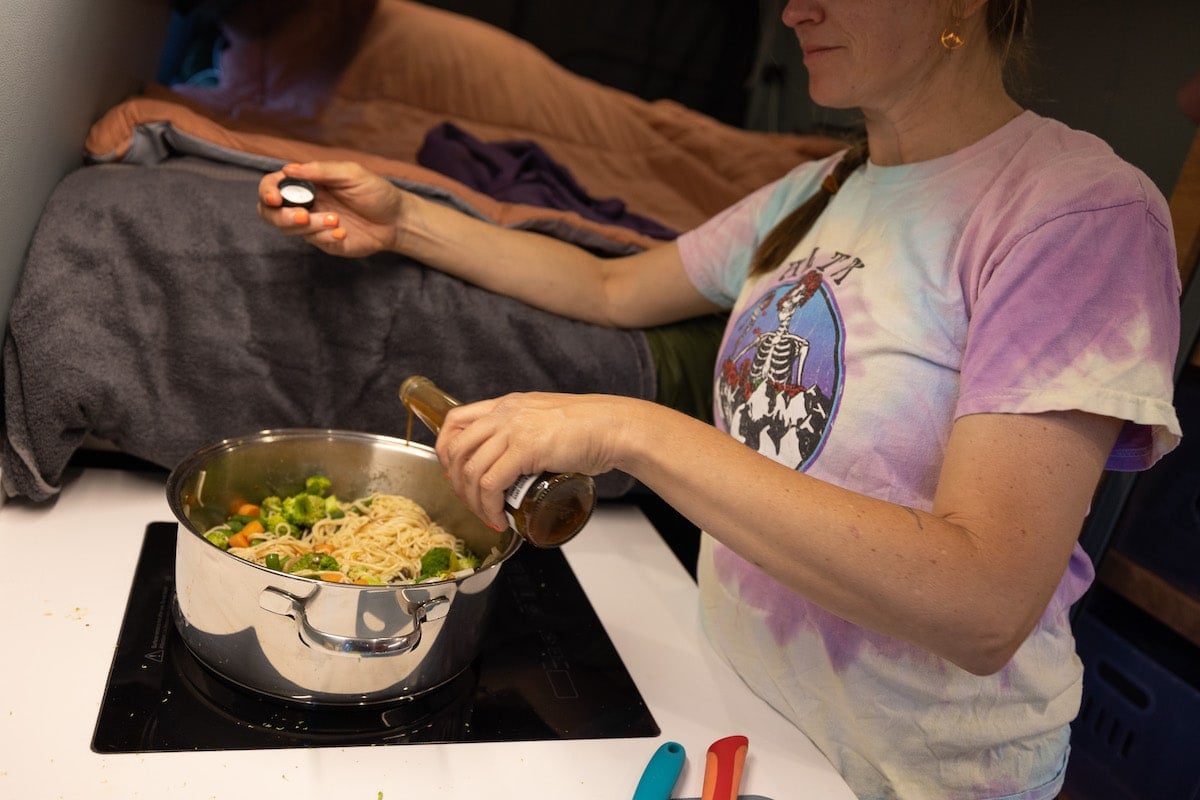 Bearfoot Theory founder Kristen Bor dumping sauce into a pan in a camper van with noodles and vegetables inside