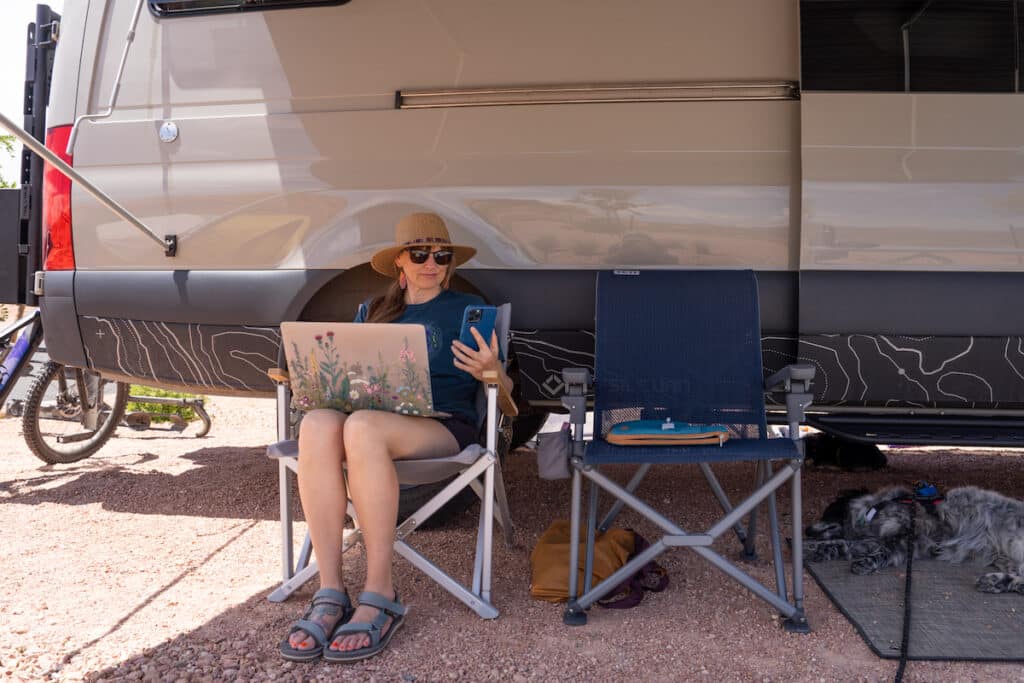 Kristen Bor with a laptop and her phone in her hand sitting in a camp chair that is next to a sprinter van