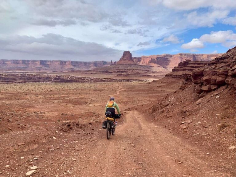 Female bikepacker on fully loaded bike pedaling down red dirt road in Utah with red mesas and bluffs in distance