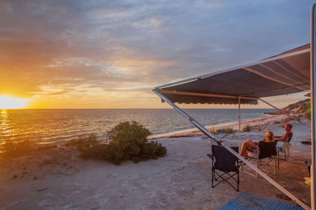 Man and woman relaxing in chairs under campervan awning on beach at sunset