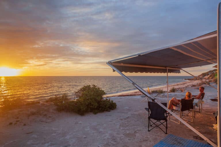 Man and woman relaxing in chairs under campervan awning on beach at sunset