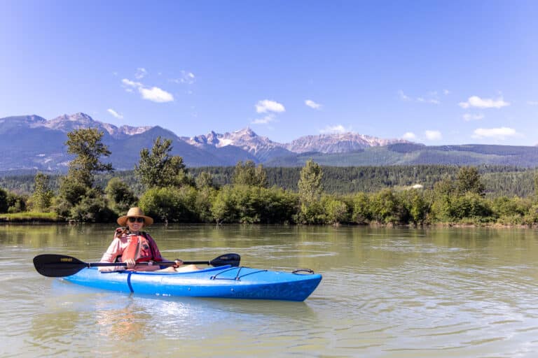 A woman smiles at the camera from her blue kayak. She is paddling in Golden Columbia River in Golden BC. She is wearing a life vest and sun hat while holding her paddle and not paddling.