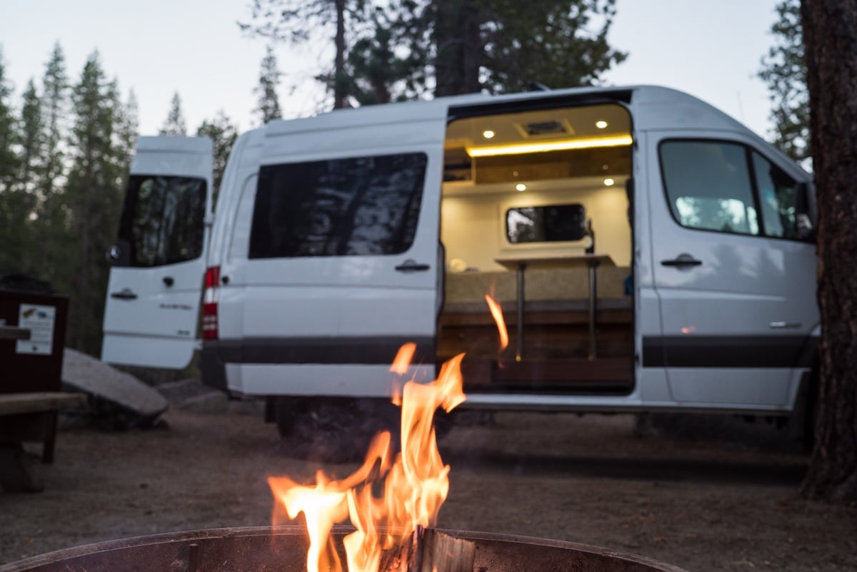 White Sprinter Van parked in front of a campsite with a fire