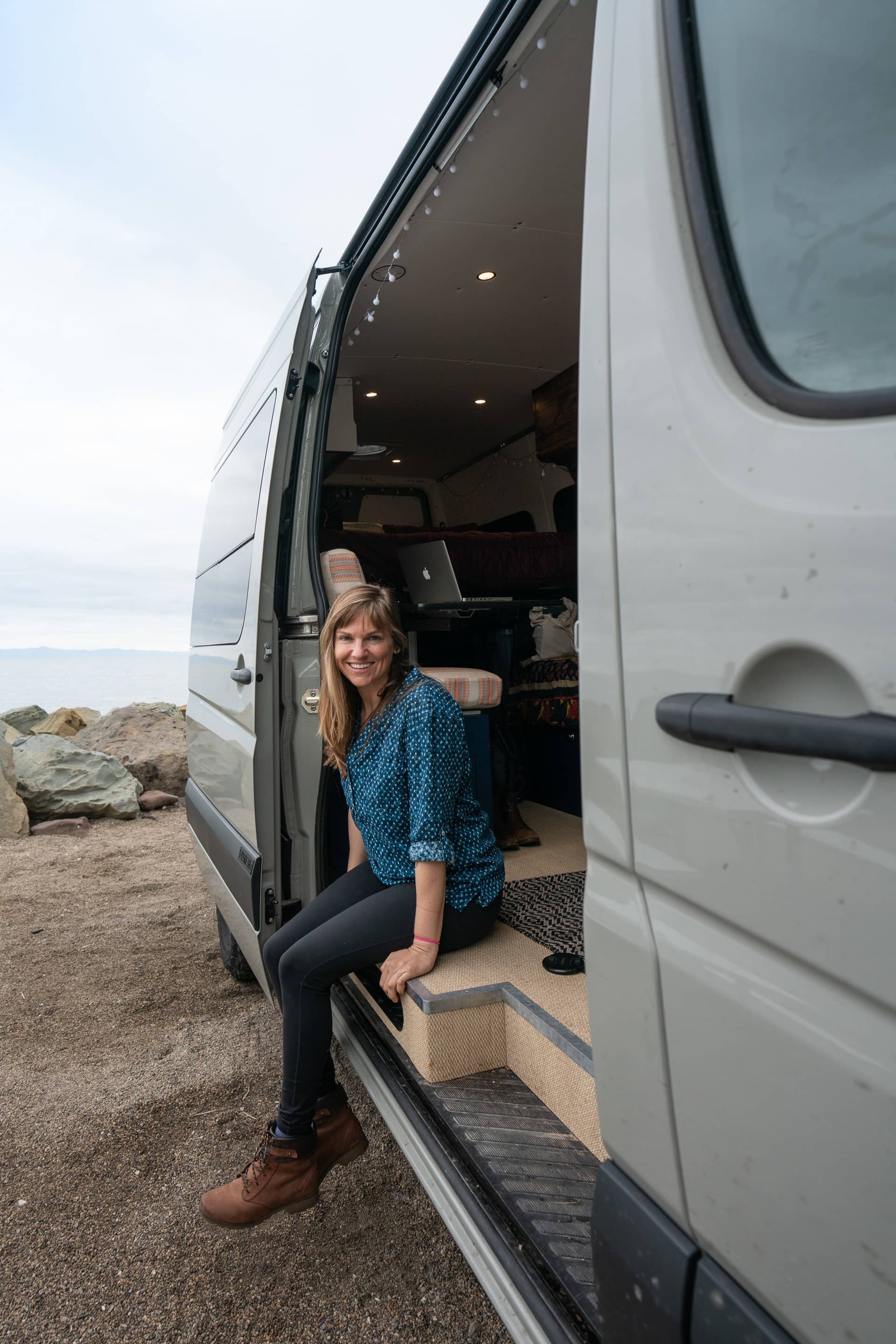A woman sitting on the edge of her Sprinter side door wearing Carve Designs Dylan Gauze Shirt (top) & Prana Transform High Waisted Leggings (bottom)