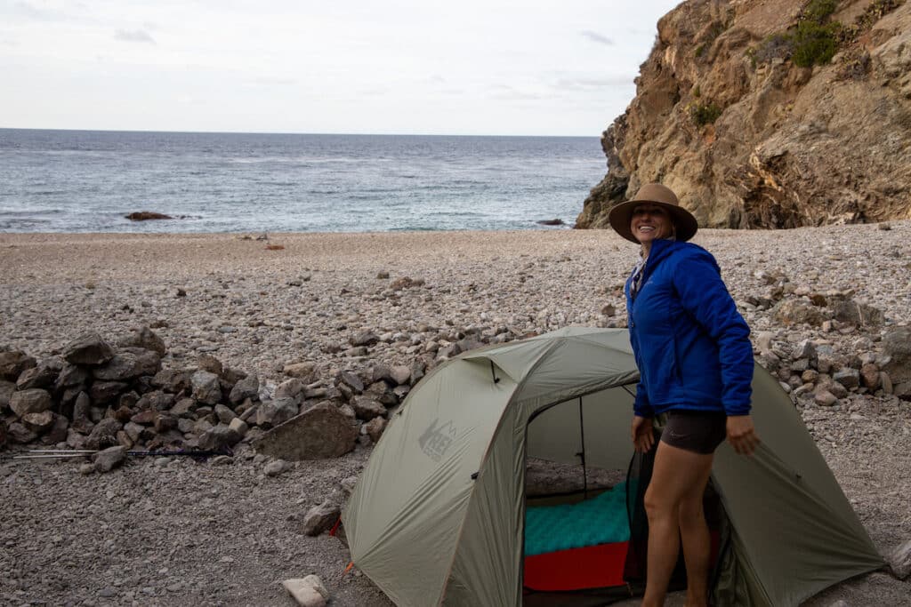 A woman smiles at the camera next to her REI Quarter Dome 2 backpacking tent. She's wearing a blue Arcteryx Atom jacket. She is next to the ocean on the Trans Catalina Trail