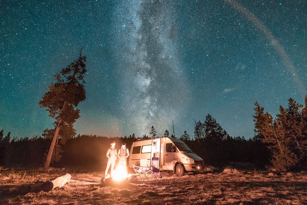 Vanlife couple standing in front of campfire and a campervan under the Milky Way at night