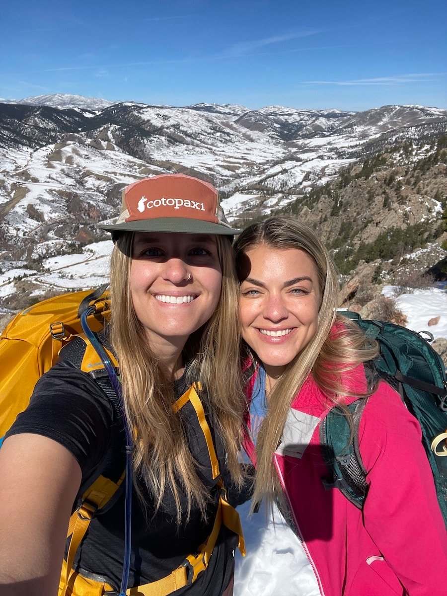 Two women smiling on a hike with snow covered hills in the background