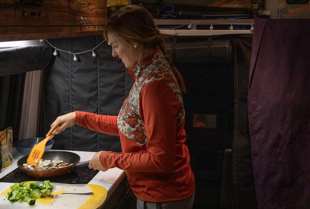 A woman cooks inside her Sprinter Van on an induction cooktop. She is sautéing vegetables and has chopped broccoli on the side.