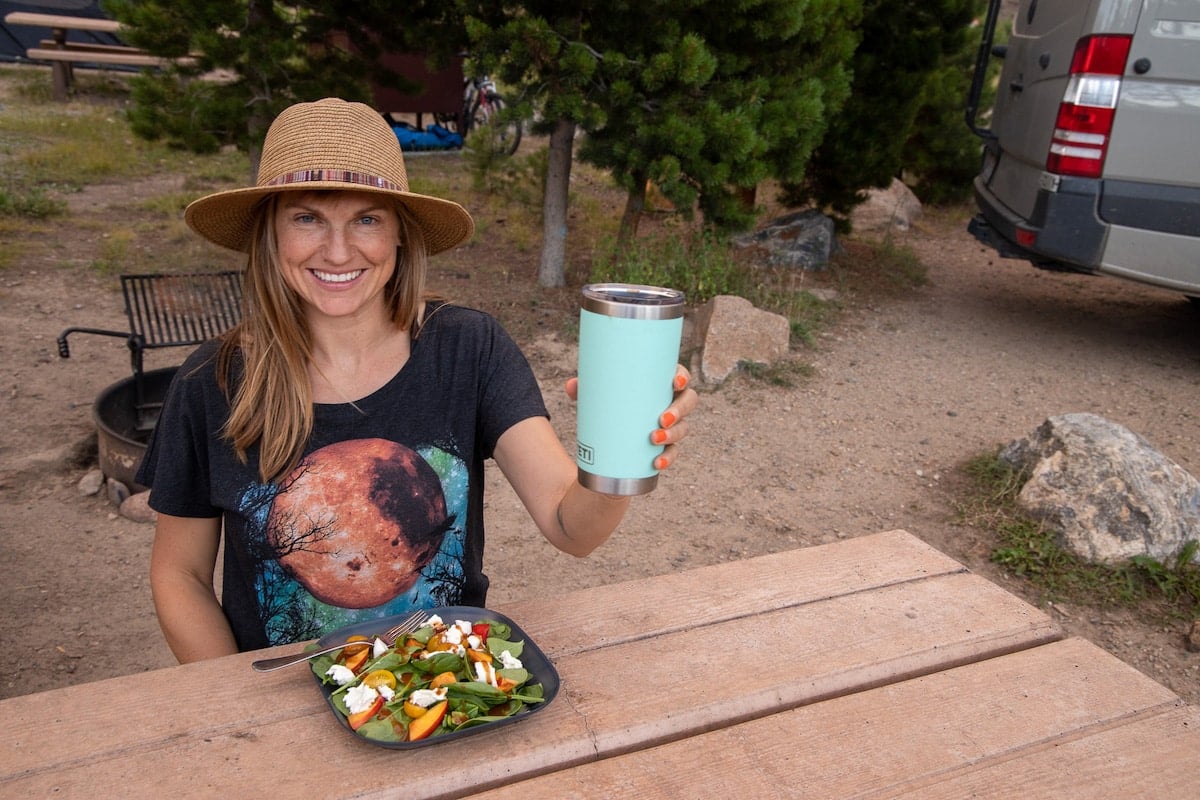 Bearfoot Theory founder Kristen Bor eating outside at a table with a plate full of vegetables and holding a blue thermos cup while smiling