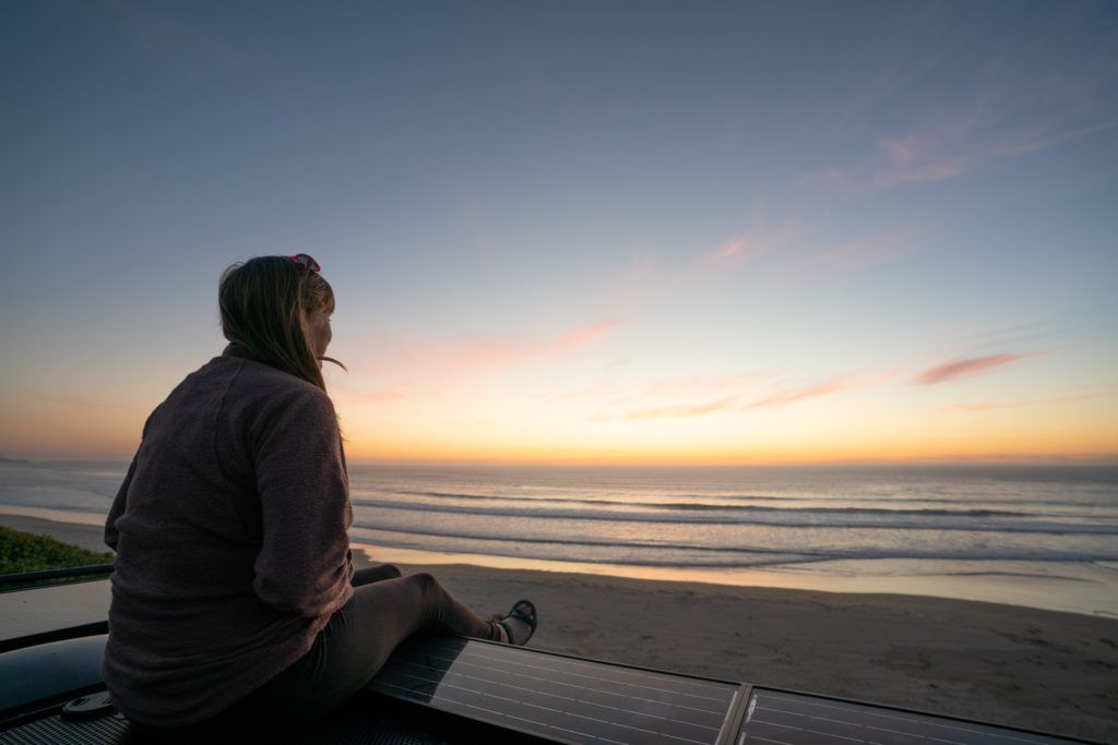 Bearfoot Theory founder Kristen Bor sitting on top of her Sprinter Van during sunset at the beach in California