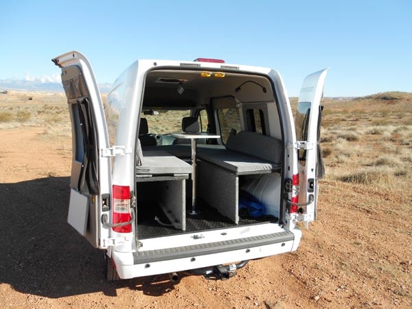 The back of a small rental campervan from Campervan North America with the doors open showing bench seating and a table. 