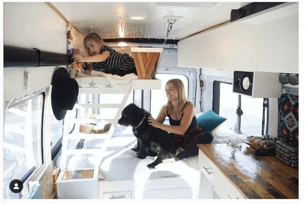 A woman sits on the bed in her campervan next to her dog with her daughter in a small bunk bed above