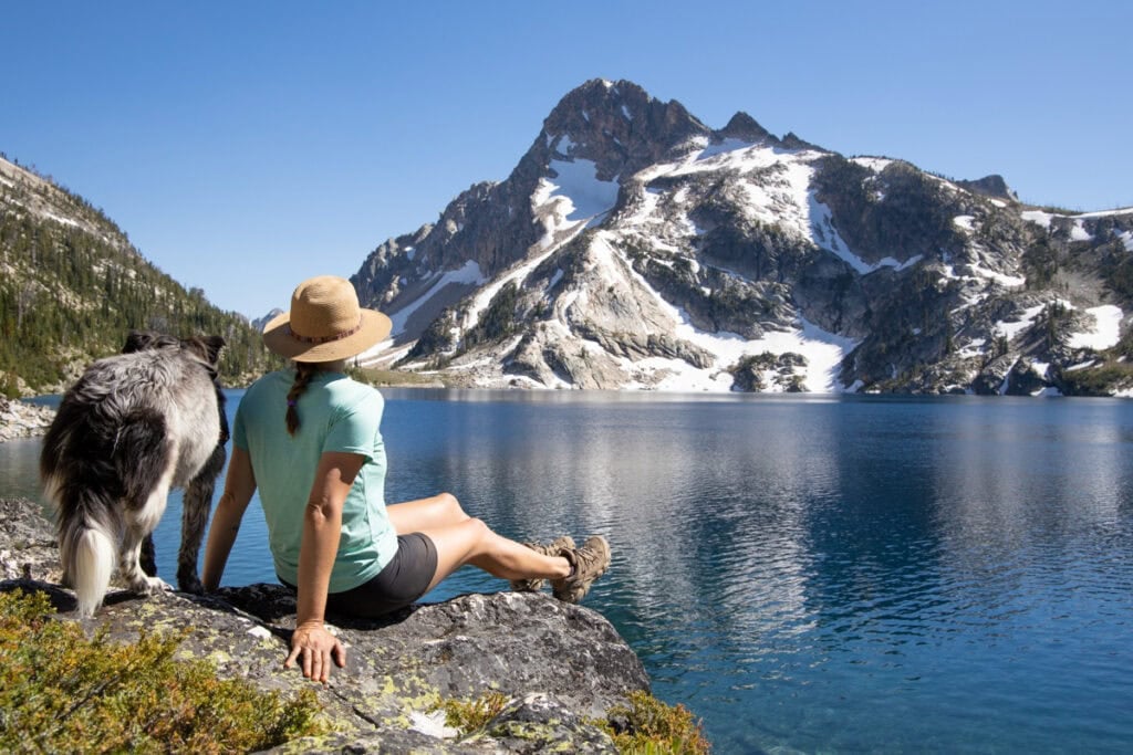 Kristen Bor sitting next to a border collie on the edge of Sawtooth Lake with snow capped mountains in the background