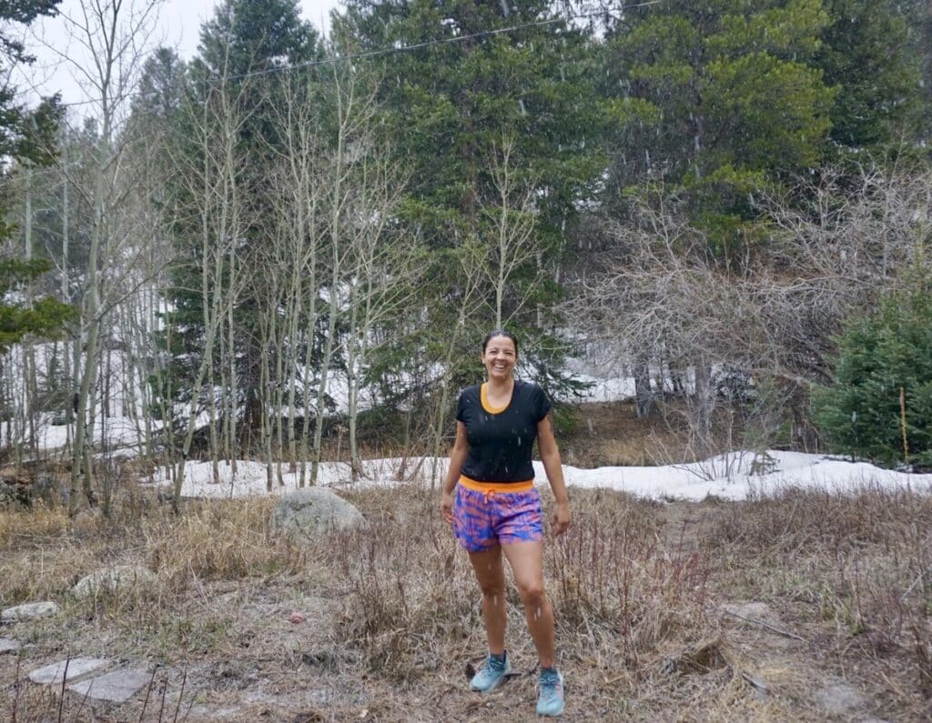 A woman smiles at the camera wearing Janji AFO 3" Shorts. There's a partially snowy trail in the background