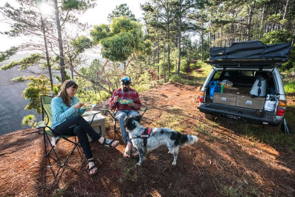 two people sitting in camp chairs eating a meal next to a truck camper