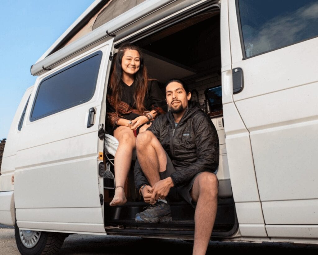 a woman and man sit in the side doorway of their white campervan