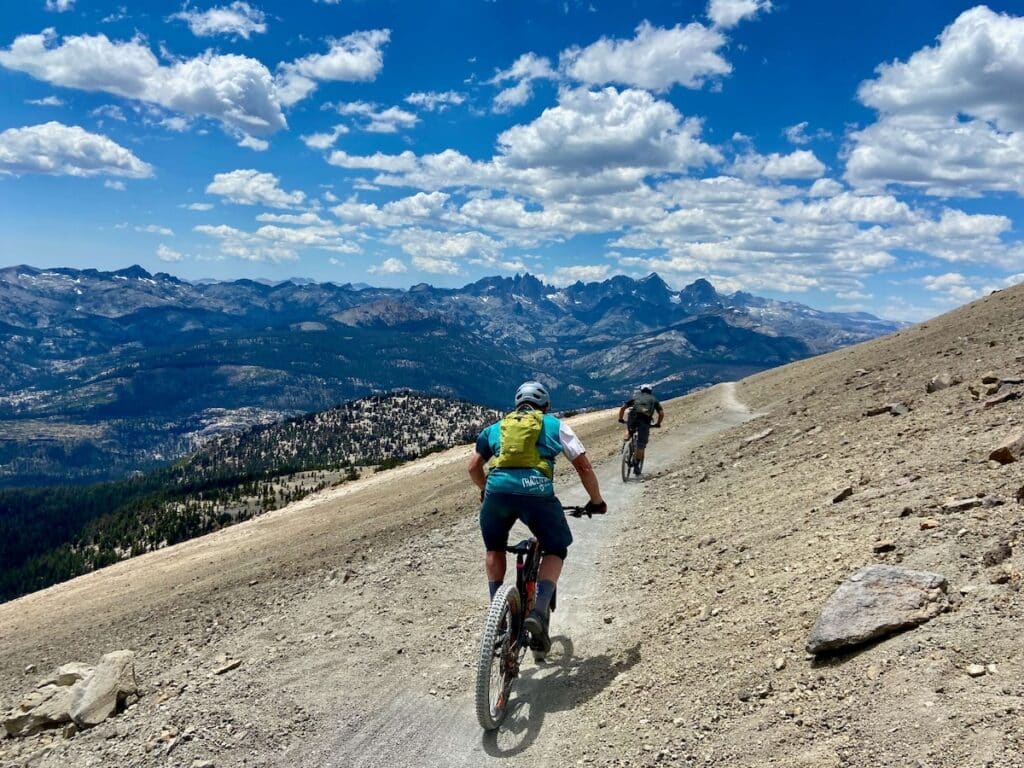 Mountain bikers riding bikes down trail from summit of Mammoth Mountain with views of mountain range in the distance