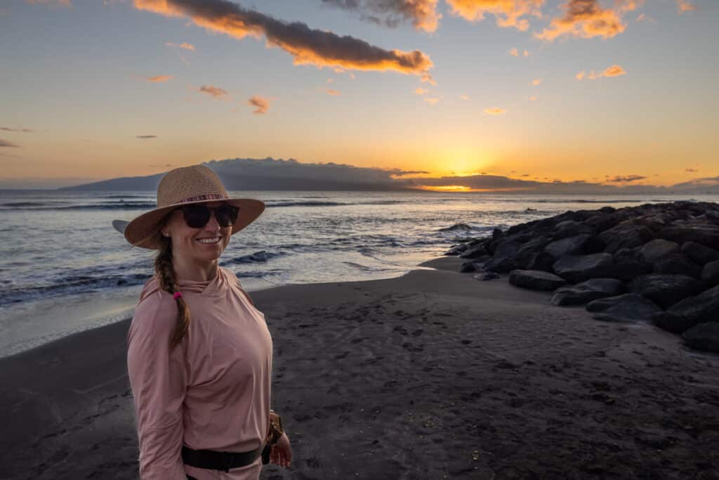 Kristen Bor standing on the beach in Maui at sunset wearing a pink REI Sahara Shade Sun Protection Hoody