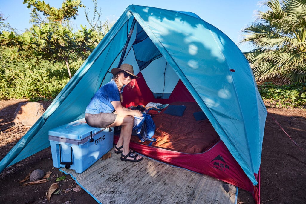 A woman sits on an RTIC 45 cooler outside her camping tent
