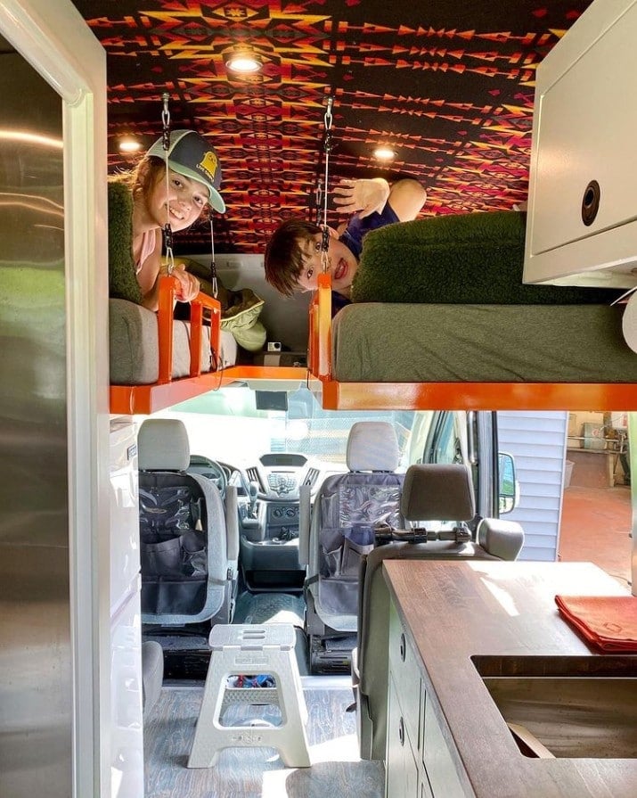 Two kids smiling in fold out beds near the cab of a family campervan