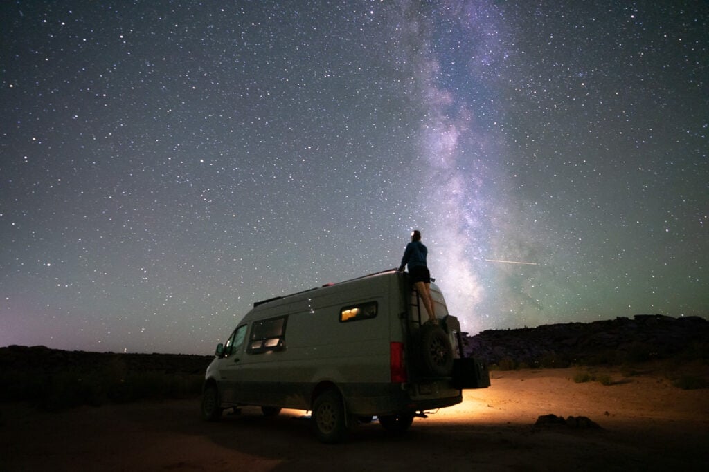 Woman standing on top of a ladder ont the back of a Sprinter camper van under a starry sky