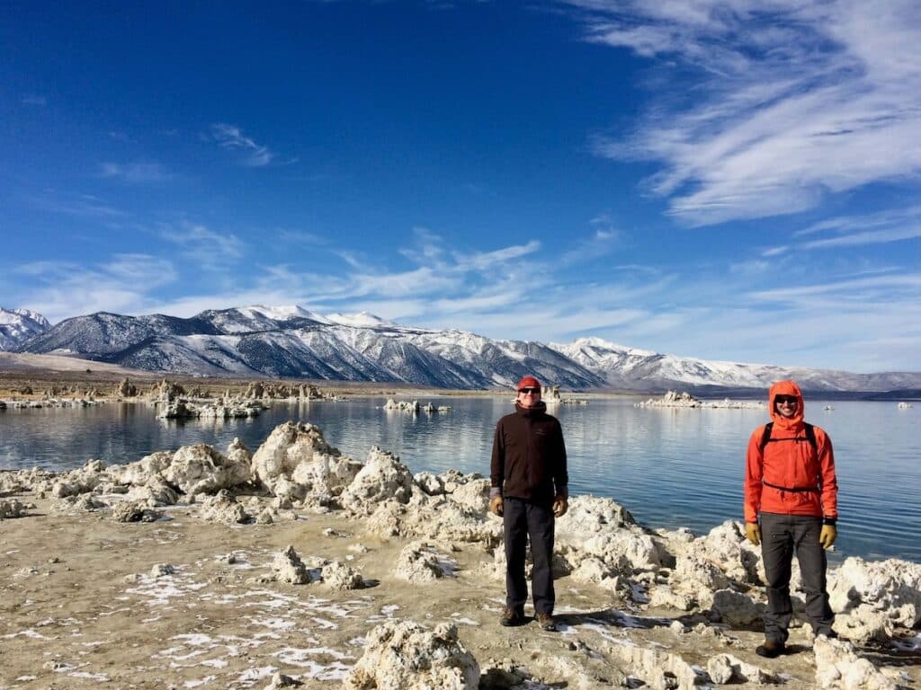 Two men dressed in warm clothes standing in front of Mono Lake in California next to limestone tufa formations and snowy mountains in the background