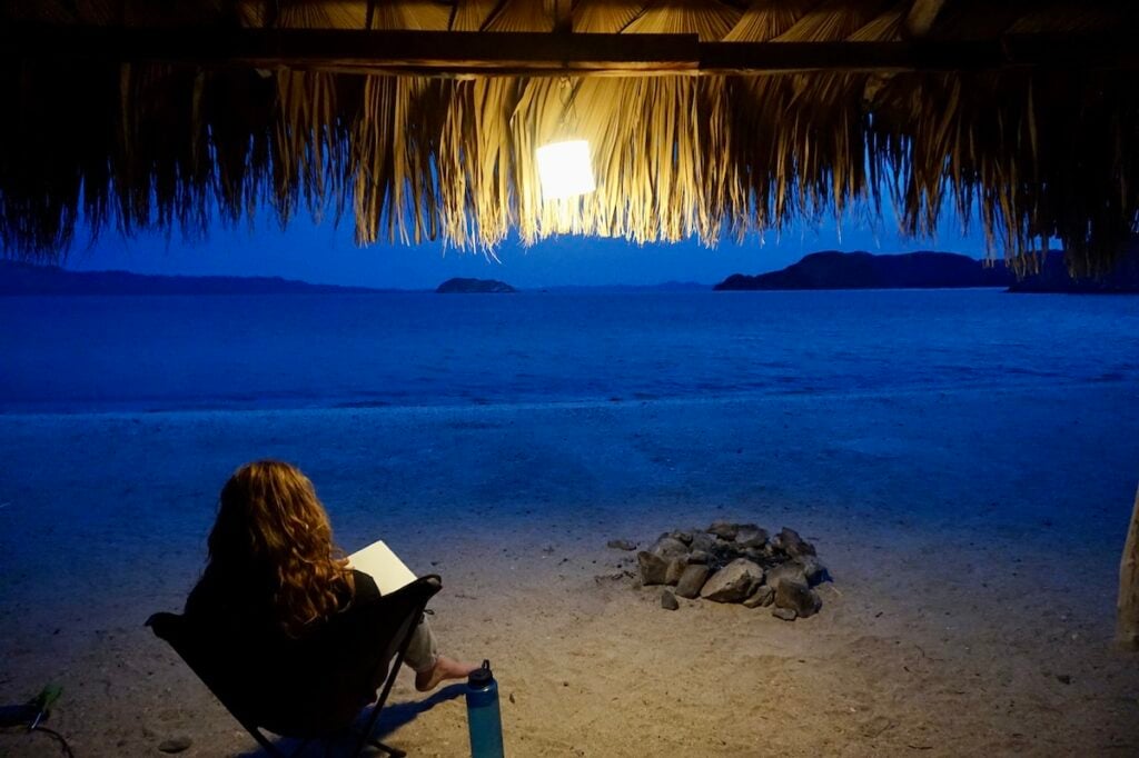 A woman journals in a palapa on the beach in Baja Mexico using the MPOWERED Luci Solar Lantern