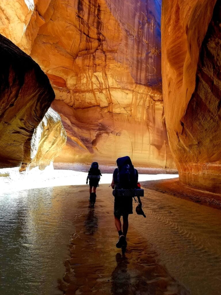 Two backpackers hiking through shallow river under tall sandstone cliff walls in Paria Canyon in Utah