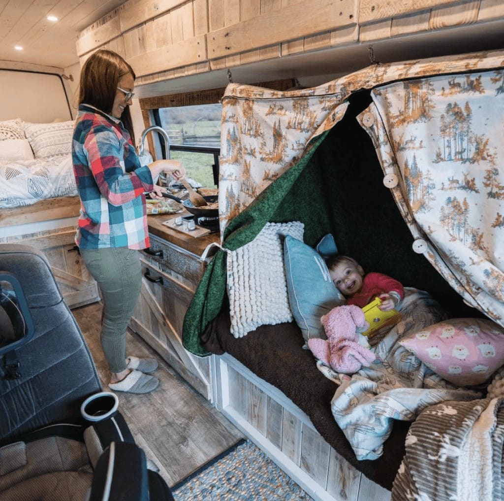 A woman cooking dinner in her campervan with a toddler in a small bed in front