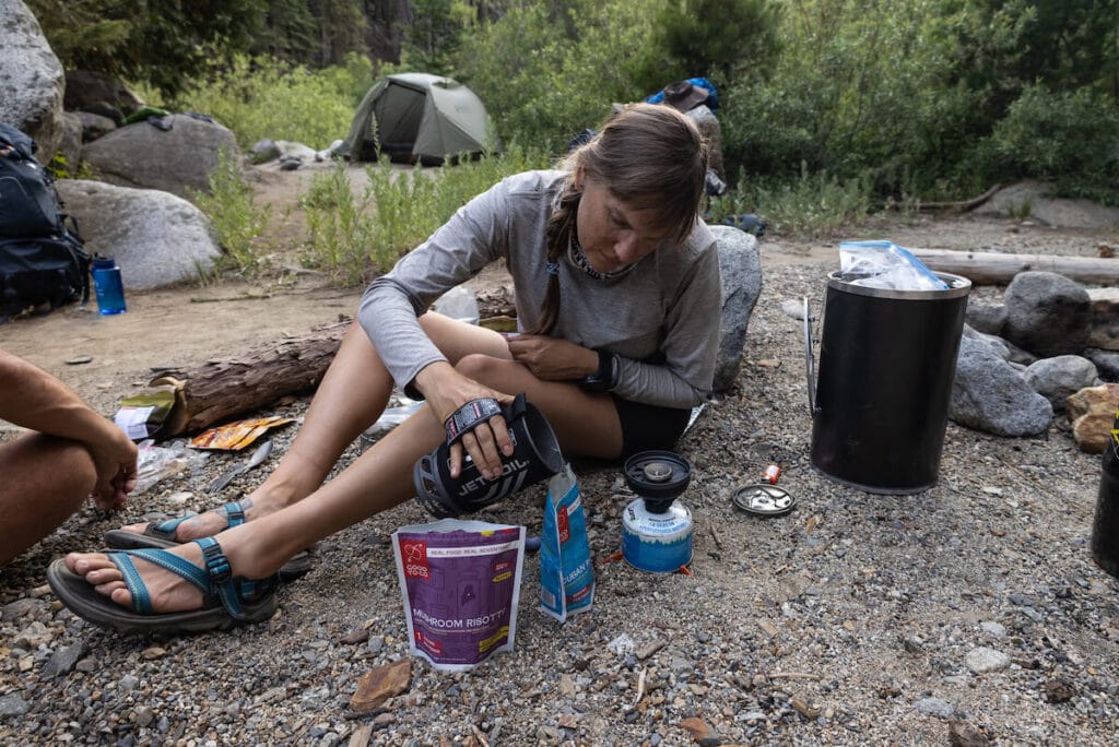 Kristen pouring hot water from Jet Boil backpacking stove into dehydrated meal pouch on a backpacking trip with backpacking gear spread out around her