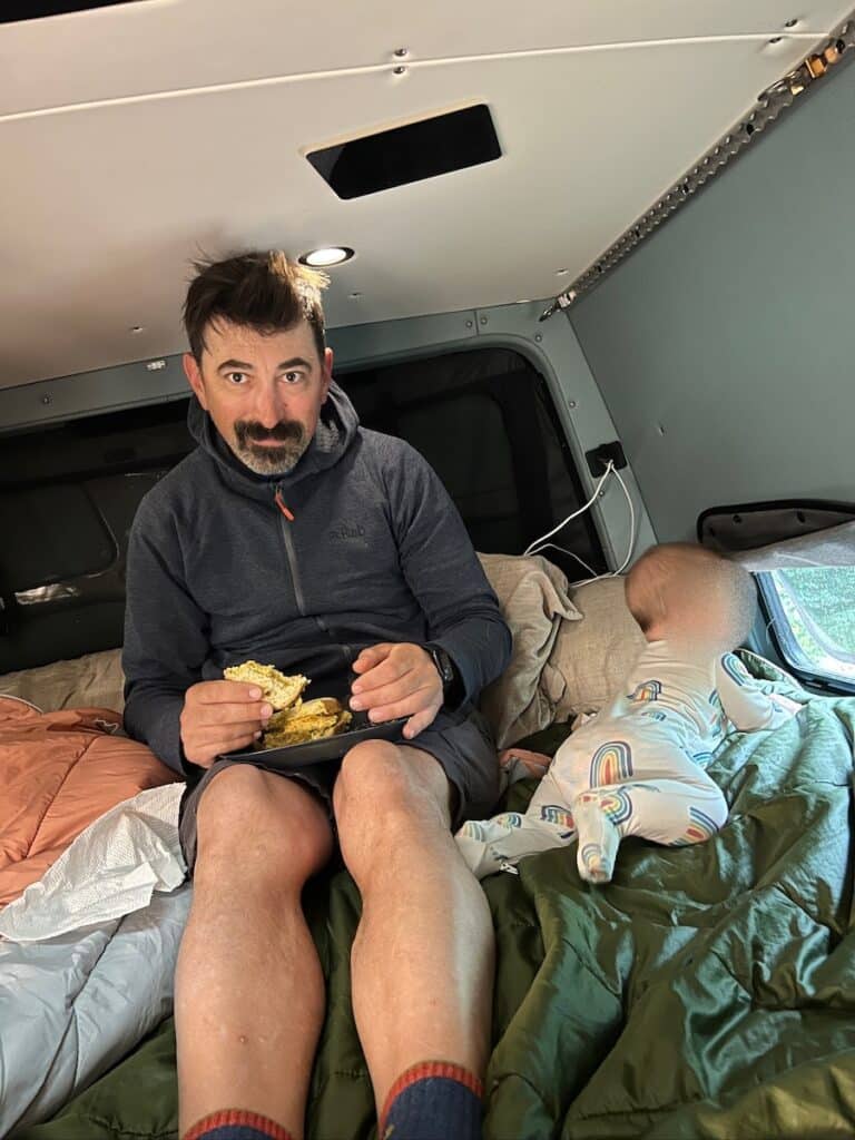 Man sitting in a Sprinter Van bed next to his baby eating a sandwich
