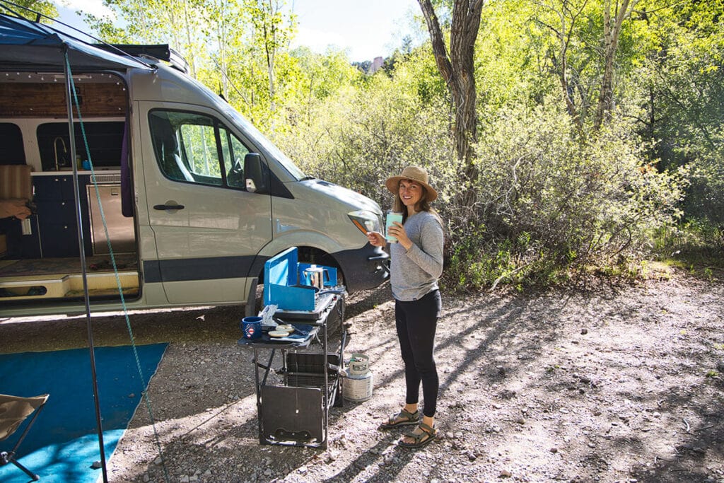 A woman stands next to the Eureka Ignite Camp Stove sitting on a GCI Outdoor Slim Fold Cook Station table. She is standing next to her Sprinter van.