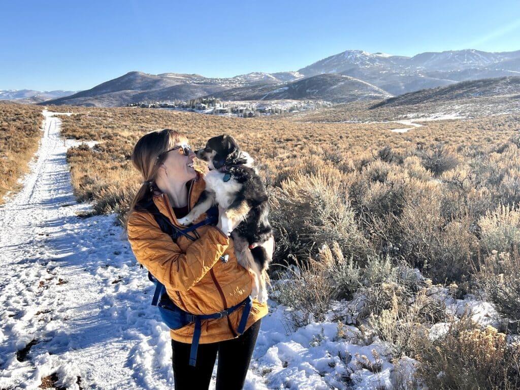 BFT Founder Kristen smiling in a golden yellow Patagonia Nano Puff on a snowy hike in Utah while snuggling her dog