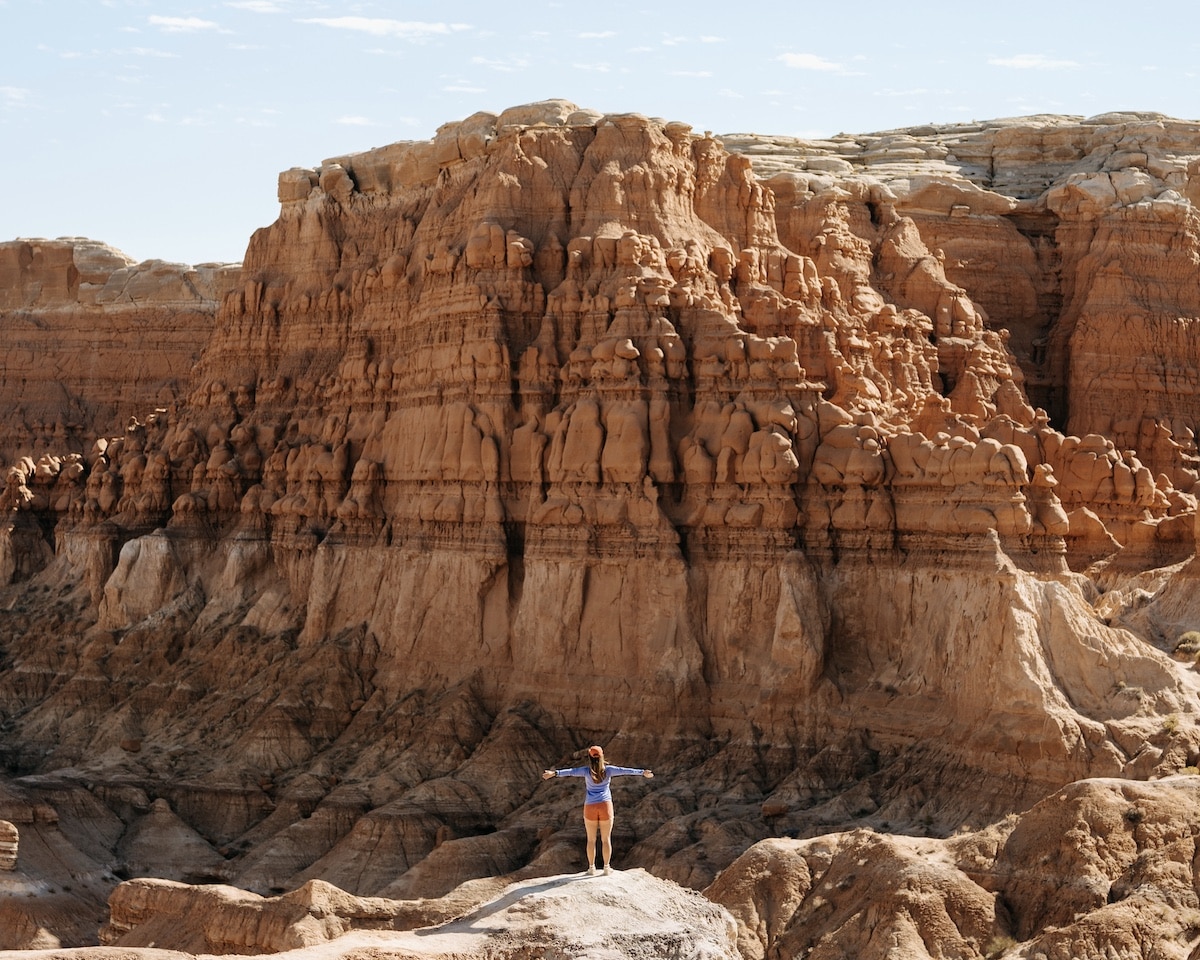 Female hiker with arms spread wide facing an orange rock formation on the Goblin's Lair trail in Goblin Valley State Park in Utah