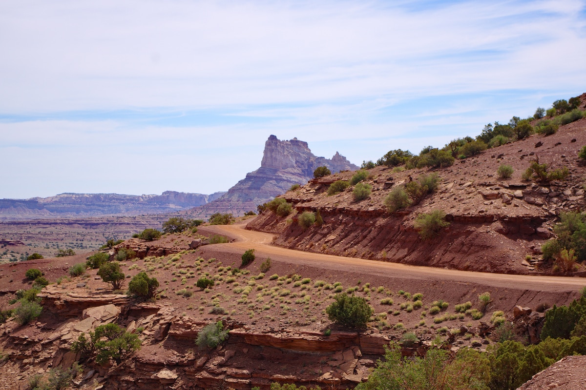 A view of Temple Mountain Road with an orange dirt road, green shrubs, and rock formations in the distance outside of Goblin Valley State Park in Utah