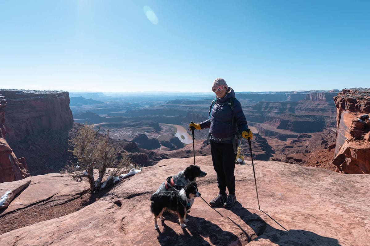 Kristen Bor with hiking poles and dogs standing at Dead Horse State Park lookout over Colorado River and Canyonlands National Park in Utah