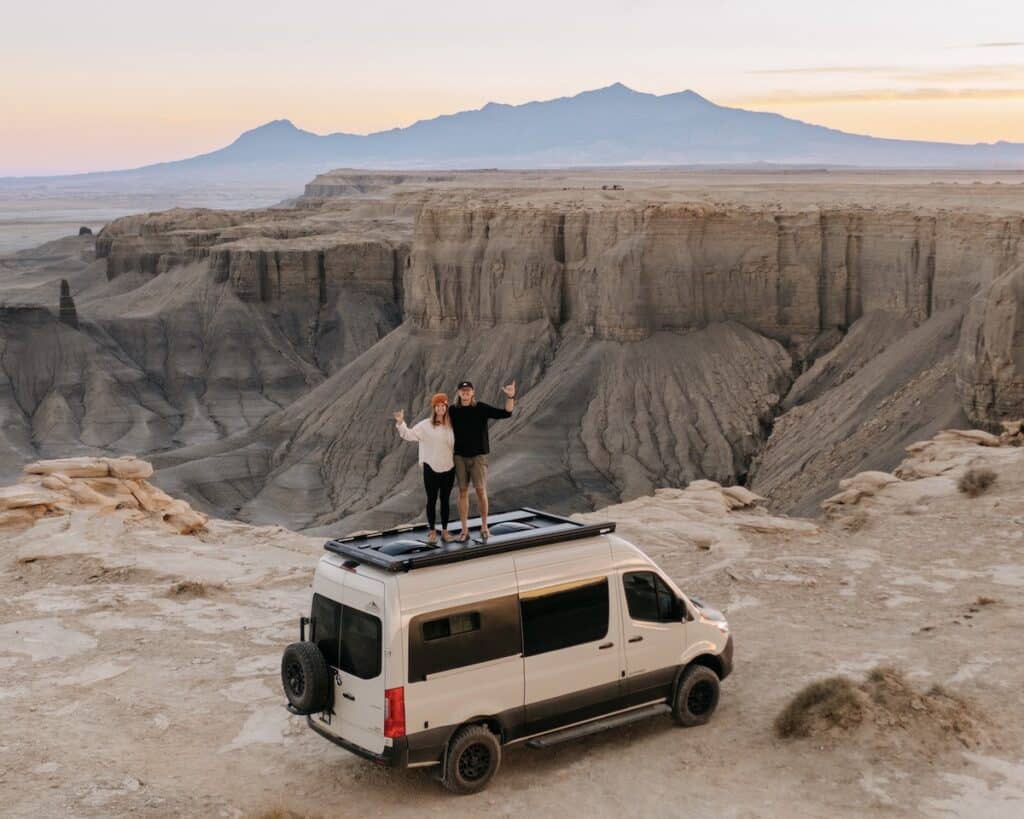 A man and a woman standing on top of a pebble gray Sprinter van at Moonscape Overlook near Hanksville, Utah