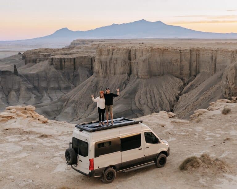 A man and a woman standing on top of a pebble gray Sprinter van at Moonscape Overlook near Hanksville, Utah