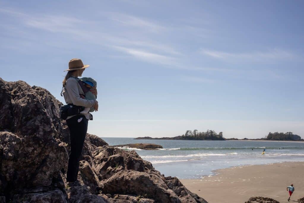 Woman carrying baby strapped to her chest standing on rocky overlook out onto the ocean and beach