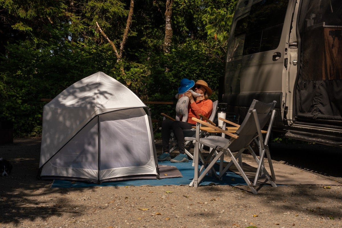 A woman holds her baby sitting next to the Veer Basecamp Shelter. The baby is wearing the Sunday Afternoons Baby SunSprout Hat while camping