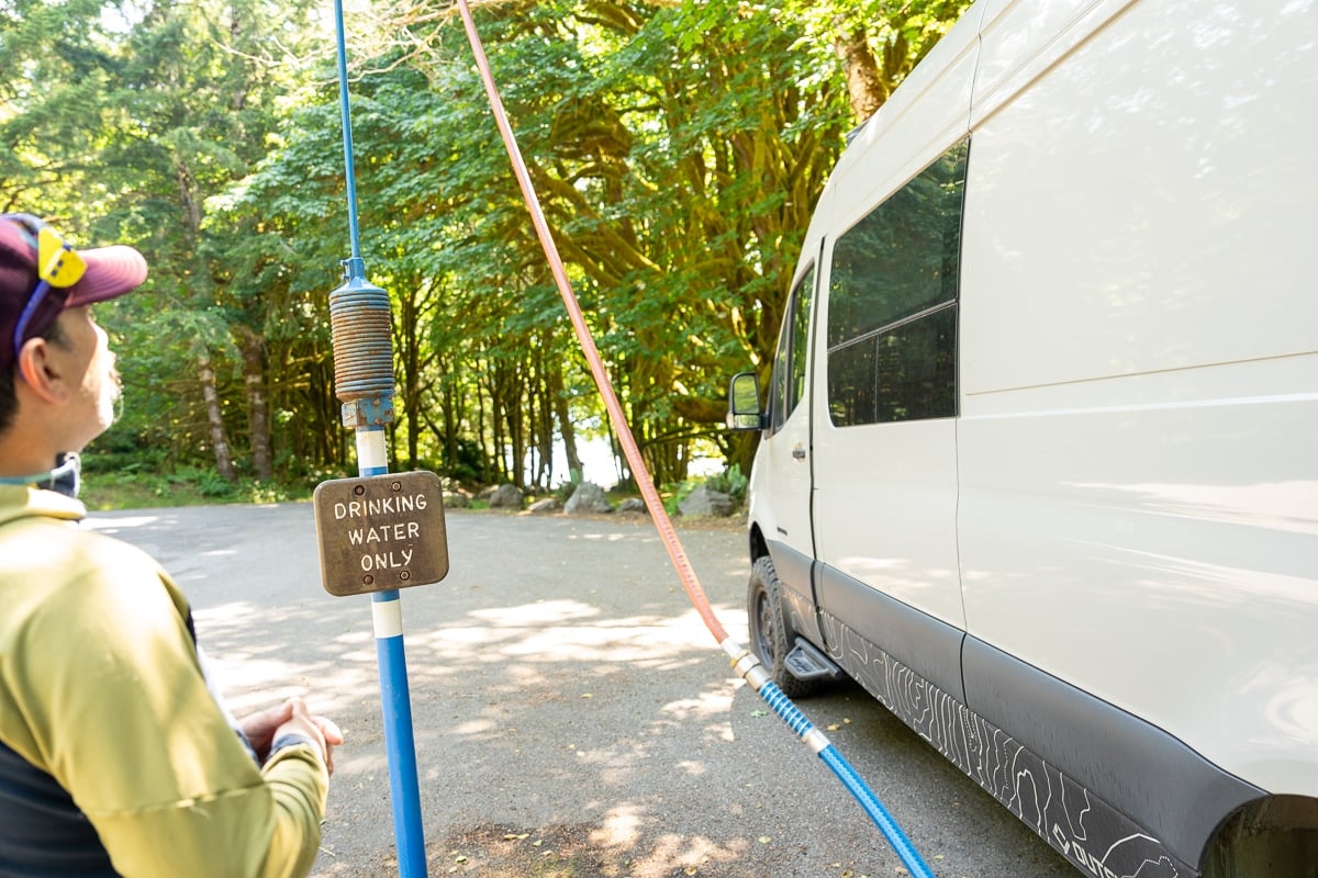Man standing next to potable water spigot filing up the tanks in a Sprinter Van with a hose