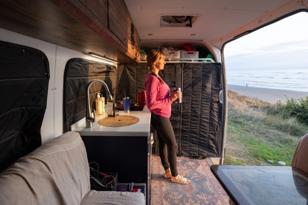 Bearfoot Theory founder Kristen Bor standing in the door of her Sprinter Camper Van while drinking tea during sunset looking out at the ocean